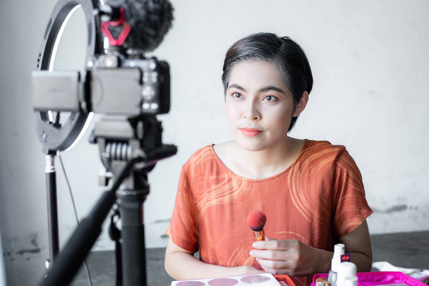 Ασιατική όμορφη blogger ομορφιά της μόδας κοιτάζει σε μια ψηφιακή φωτογραφική μηχανή. κριτικές προϊόν ομορφιάς για video blog, μιλώντας σε καλλυντικά κρατώντας μια παλέτα μακιγιάζ ενώ ηχογραφεί το βίντεό της. - Φωτογραφία, εικόνα