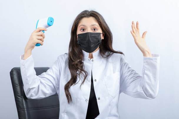 Portrait de femme médecin en masque médical et manteau blanc tenant thermomètre. Photo de haute qualité - Photo, image