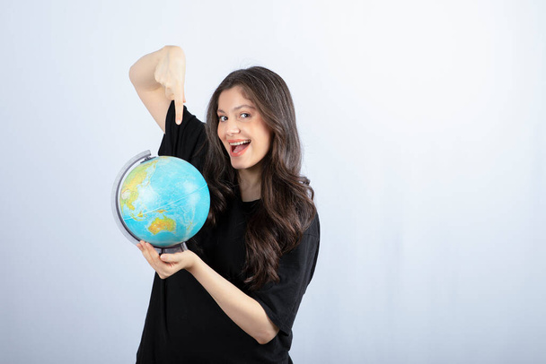 Morena sorrindo mulher com cabelos longos apontando para o globo mundial e posando. Foto de alta qualidade - Foto, Imagem