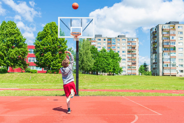 Aantrekkelijke jonge jongen schieten bal naar de hoepel op speeltuin.schattig jonge jongen speelt basketbal op de speelplaats in de zomer warme dag. - Foto, afbeelding