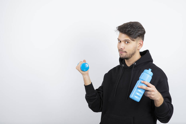 Portrait of young handsome guy holding blue dumbbell and water bottle. High quality photo - 写真・画像