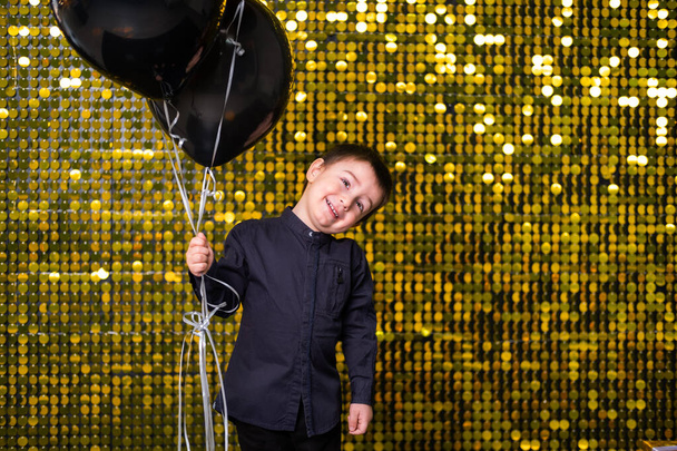 child boy holding black balloons on background with gold shiny sequins, paillettes - Photo, Image