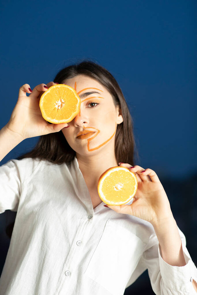 Εικόνα της νεαρής γυναίκας με ζωγραφισμένο πρόσωπο που κρατά μισό πορτοκάλι. Υψηλής ποιότητας φωτογραφία  - Φωτογραφία, εικόνα