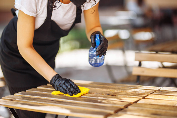 Disinfection of a cafe, restaurant, coffee shop before opening. A waitress wearing a protective mask and gloves decontaminates tables and chairs - Photo, Image