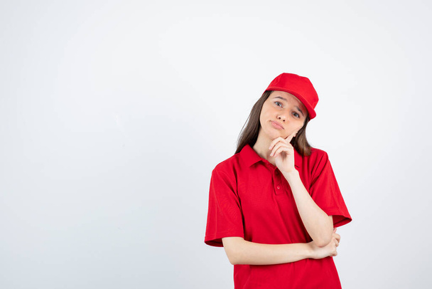 Photo of young female courier in red uniform thinking over white. High quality photo - Photo, Image