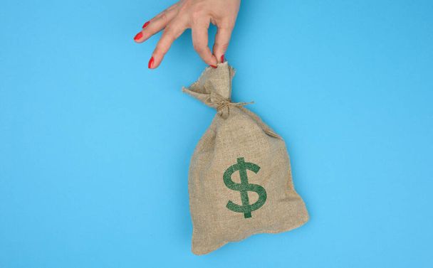 mão segura um saco de lona cheio de dinheiro com um ícone de dólar americano em um fundo azul. Conceito de planeamento orçamental, lucros e poupanças, resultado financeiro final - Foto, Imagem