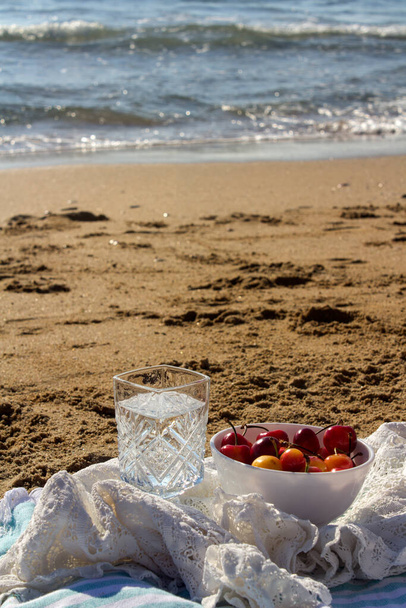 Пікнік на пляжі, миска з солодкою вишнею, склянка води на піску. Спокійне блакитне море на фоні. Фото літнього дня
.  - Фото, зображення