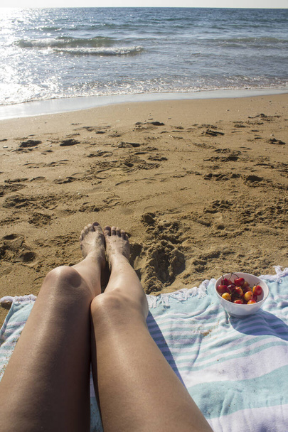 黄金の砂の上の女性の足。女性の足トップビューの写真。ビーチで若い女性。夏の脱出だ。海での休暇。ターコイズブルーのビーチタオルの上の足。ビーチで夏の楽しみ。女は太陽の下で休んでいる。日光浴をする女.  - 写真・画像