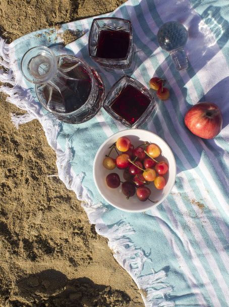 Gyönyörű tengerparti táj. Piknik a parton. vörösbor, két pohár és cseresznyés tál. homok és nyugodt tengervíz a háttérben. Nyári hétvége.  - Fotó, kép