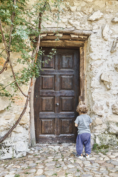 Πίσω όψη ενός μοναχικού μικρού παιδιού που στέκεται έξω μπροστά από την κλειστή ξύλινη πόρτα ενός παλιού σπιτιού. Έννοια της μοναξιάς. Κάθετη φωτογραφία. - Φωτογραφία, εικόνα