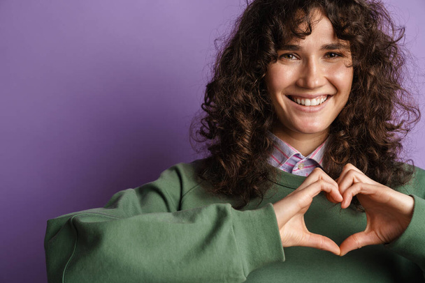 Happy beautiful curly girl smiling and gesturing heart sign with fingers isolated over purple background - Fotó, kép