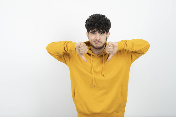 Photo of a young brunette man in yellow hoodie standing and showing thumbs down . High quality photo - Photo, image