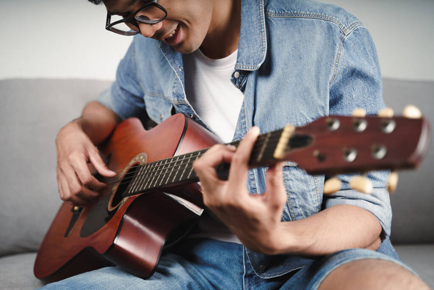 Nauti komea aasialainen mies harjoitellaan tai soittaa kitaraa sohvalla olohuoneessa  - Valokuva, kuva