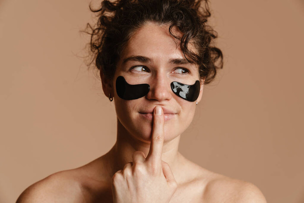 Half-naked curly woman with eye patches showing silence gesture isolated over beige background - Photo, Image