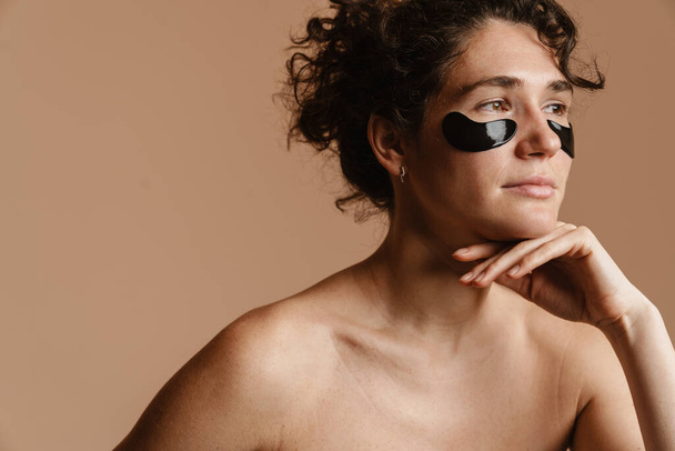 Half-naked woman looking aside while posing with eye patches isolated over beige background - Фото, зображення