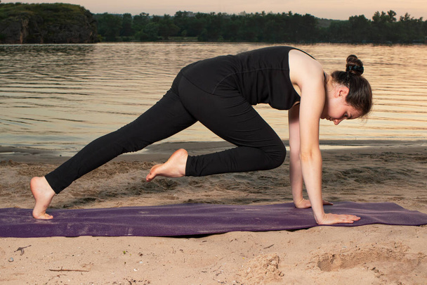 A woman in black sportswear does yoga on the beach in the evening. Pose plank with a raised leg, exercises for the press. - Photo, Image