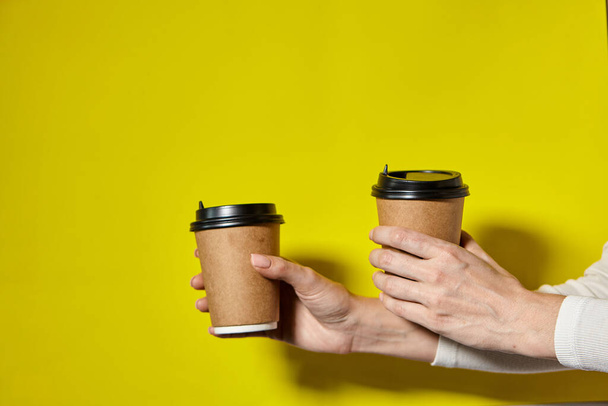 手は黒の蓋で2つのカップ茶色の紙を保持.2つのコーヒー特別オファーまたはプロモーション。黄色の画面の背景に2つのカップを保持手。紅茶やコーヒーを移動します。黒蓋の茶色い紙コップ. - 写真・画像