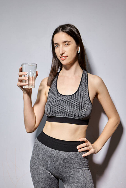 Eine Frau in Sportkleidung und mit drahtlosen Kopfhörern trinkt Wasser. das Konzept eines gesunden Lebensstils und sportlicher Aktivitäten - Foto, Bild