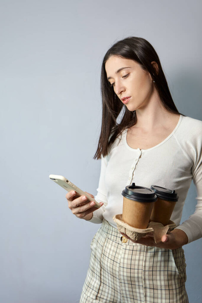 Kobieta w białej bluzie trzyma dwie filiżanki kawy w jednej ręce, a smartfona w drugiej. Herbata lub kawa do zabrania. Komunikacja zdalna - Zdjęcie, obraz