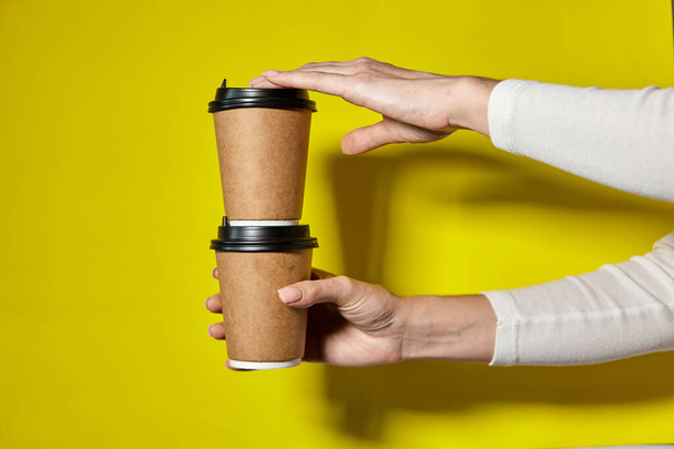Hands holding two cups brown paper with black lid. Two coffee special offer or promo. Hands holding two cups on yellow screen background. Tea or Coffee to go. Brown paper cup with black lid. - Zdjęcie, obraz
