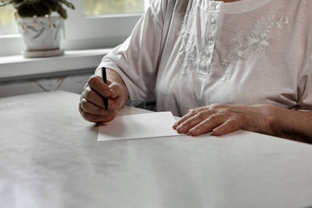Una donna anziana scrive a casa su un pezzo di carta. Interessi di pensionati. Comunicazione remota, invio di email. Il processo di scrittura di una nota - Foto, immagini
