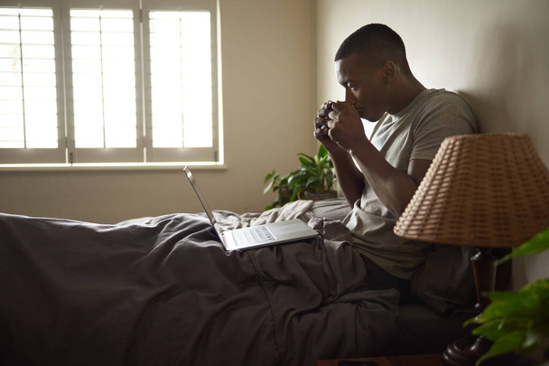 Young African man sitting in bed in the morning browsing the internet with a laptop and drinking a cup of coffee - Valokuva, kuva