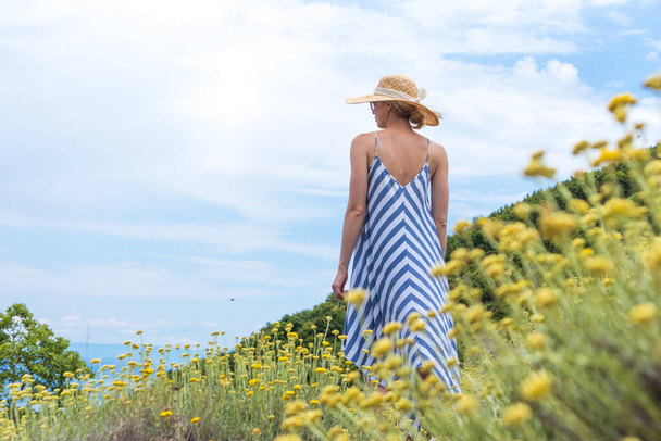 Νεαρή γυναίκα με ριγέ καλοκαιρινό φόρεμα και ψάθινο καπέλο στέκεται σε σούπερ άνθιση αγριολούλουδων, χαλαρώνοντας απολαμβάνοντας την όμορφη φύση της Αδριατικής θαλάσσιας παράκτιας φύσης της Κροατίας. - Φωτογραφία, εικόνα