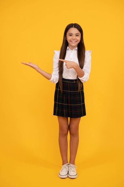 Okul üniformalı neşeli genç kız sarı arka planı işaret ediyor, duyuru. - Fotoğraf, Görsel