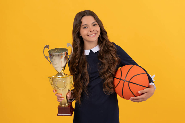 χαρούμενο κορίτσι έφηβος κατέχουν μπάλα μπάσκετ και κύπελλο πρωταθλητής στο κίτρινο φόντο, πρωταθλητής - Φωτογραφία, εικόνα
