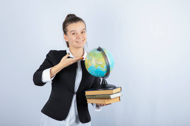 Zdjęcie pięknej młodej nauczycielki z książkami i globusem stojącej na białym tle. Wysokiej jakości zdjęcie - Zdjęcie, obraz