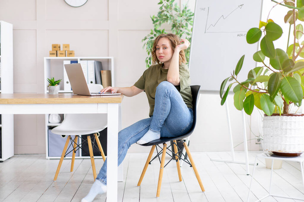 Business woman using laptop sitting desk white office interior with houseplant looking Business people Business person Online, Young and successful Dresed green shirt blue jeans barefoot relaxing  - Zdjęcie, obraz