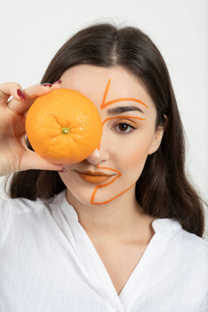 Obraz młodej kobiety z pomalowaną twarzą zakrywającą oko całymi pomarańczowymi owocami. Wysokiej jakości zdjęcie - Zdjęcie, obraz