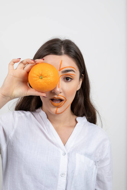 Egy fiatal nő képe festett arccal, aki narancssárga gyümölcsökkel takarta el a szemét. Kiváló minőségű fénykép - Fotó, kép