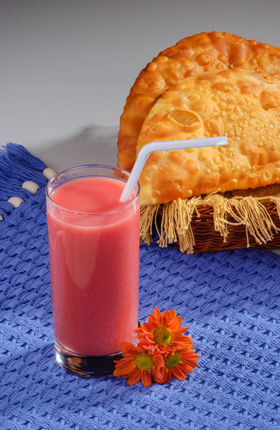 Verre de jus de fruits avec paille plastique et pastels dans un petit panier avec napperon bleu orné de fleurs rouges sur fond dégradé gris. - Photo, image