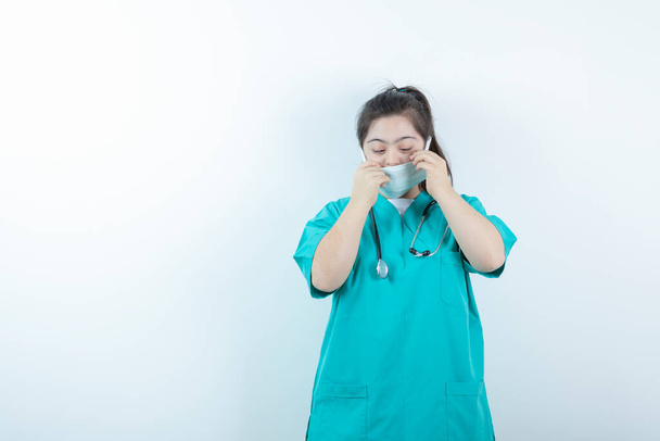 Jonge vrouwelijke verpleegster met stethoscoop met medisch gezichtsmasker tegen witte muur. Hoge kwaliteit foto - Foto, afbeelding