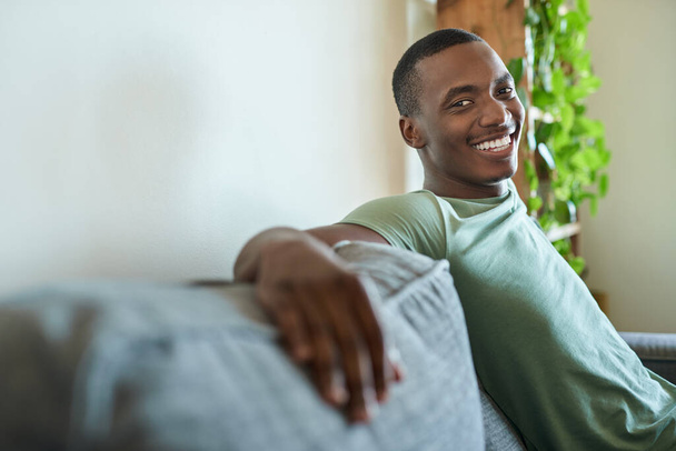 Retrato de um jovem africano sorrindo enquanto relaxa em um sofá em sua sala de estar em casa à tarde - Foto, Imagem