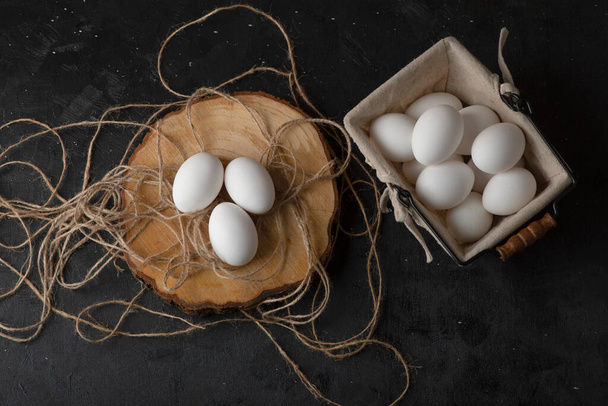 Three white raw eggs on wood piece with rope on black background. High quality photo - Photo, Image