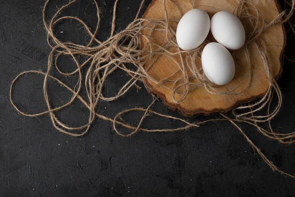 Three white raw eggs on wood piece with rope on black background. High quality photo - Foto, Imagem