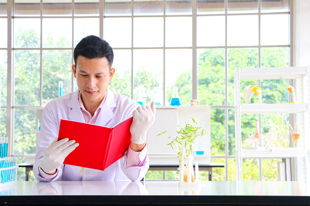 Naukowiec lub badacz w azjatyckim mężczyźnie w białym płaszczu pracuje i czyta książkę, aby znaleźć informacje. Bycie w laboratorium naukowym ze sprzętem do probówek - Zdjęcie, obraz