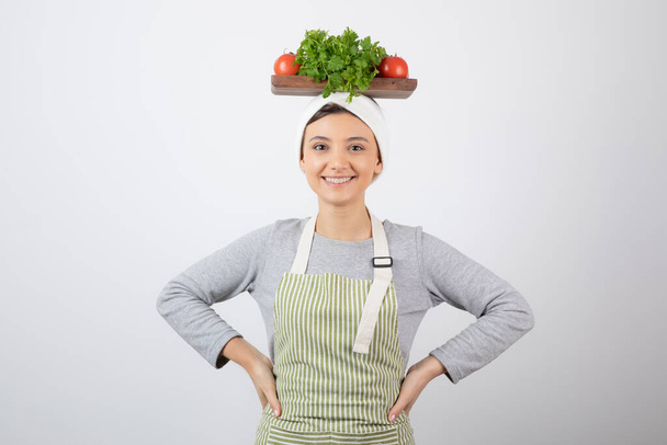 Photo of a smiling cute woman model holding a wooden board with fresh vegetables on head. High quality photo - Photo, Image