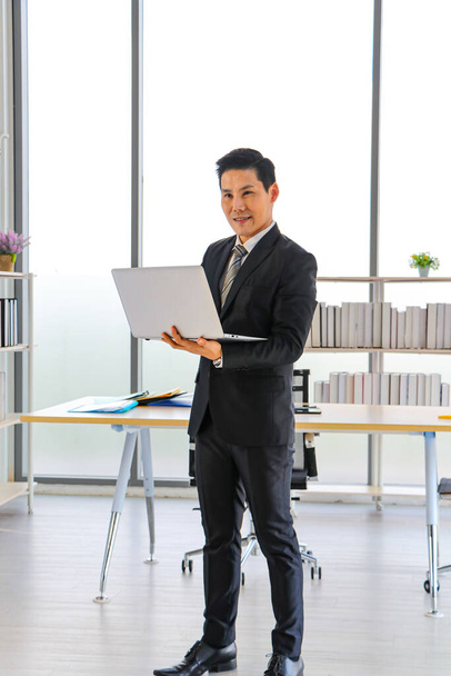 Un hombre de negocios asiático se para y sostiene una computadora portátil para trabajar en la oficina. Utiliza aplicaciones para redes sociales y noticias de tecnología en línea para estrategias empresariales. Sonríe y sé feliz - Foto, imagen