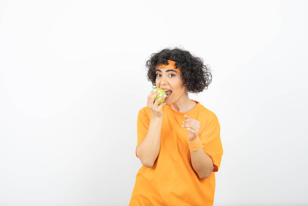 Turuncu kıyafetli, yeşil elma yiyen kıvırcık kadın. Yüksek kalite fotoğraf - Fotoğraf, Görsel
