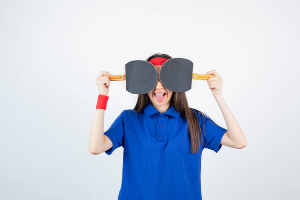 Photo of a young girl covering eyes with table tennis rackets over a white wall . High quality photo - Foto, immagini