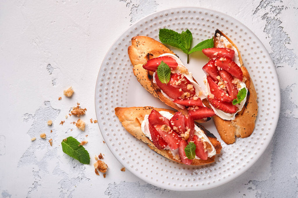 Open sandwiches with strawberries, soft cheese mint and walnut in ceramic plate on a light grey stone background. Summer and healthy dieting food, vegetarian food concept. Top view. - Photo, Image