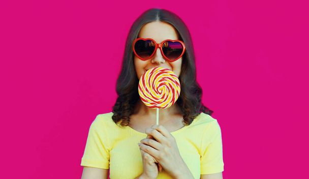 Portrait close up of young woman covering her lips with sweet colorful lollipop on a pink background - Photo, Image