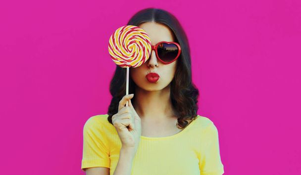 Portrait close up of cute sweet woman blowing her lips with red lipstick covering her eye with lollipop on a pink background - Zdjęcie, obraz