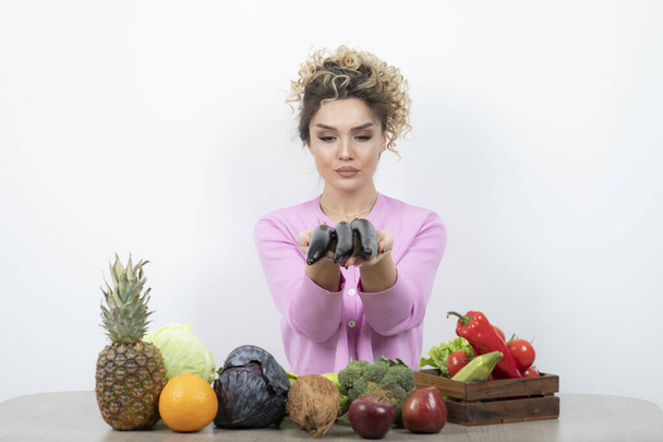 Meyve ve sebzelerle masada otururken elinde patlıcan tutan genç bir kadın. Yüksek kalite fotoğraf - Fotoğraf, Görsel