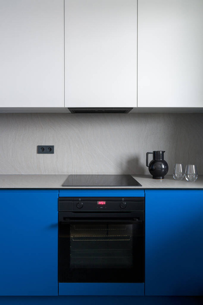 Simple black oven and induction hob in modern kitchen with white and blue cupboards and gray countertop with backsplash - Photo, image