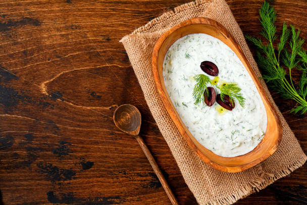 Traditional Greek sauce or dip Tzatziki, yogurt, cucumber, dill, garlic and salt oil in rustic wooden bowl on wooden background. Rustic style. Selective focus. Top view. - Photo, image