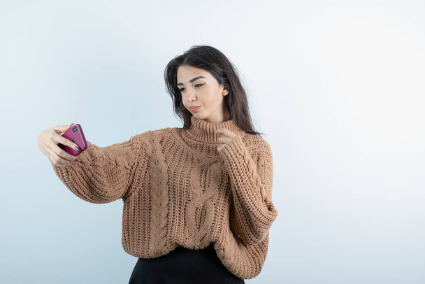 Image of beautiful woman in knitwear taking selfie on white background. High quality photo - Zdjęcie, obraz